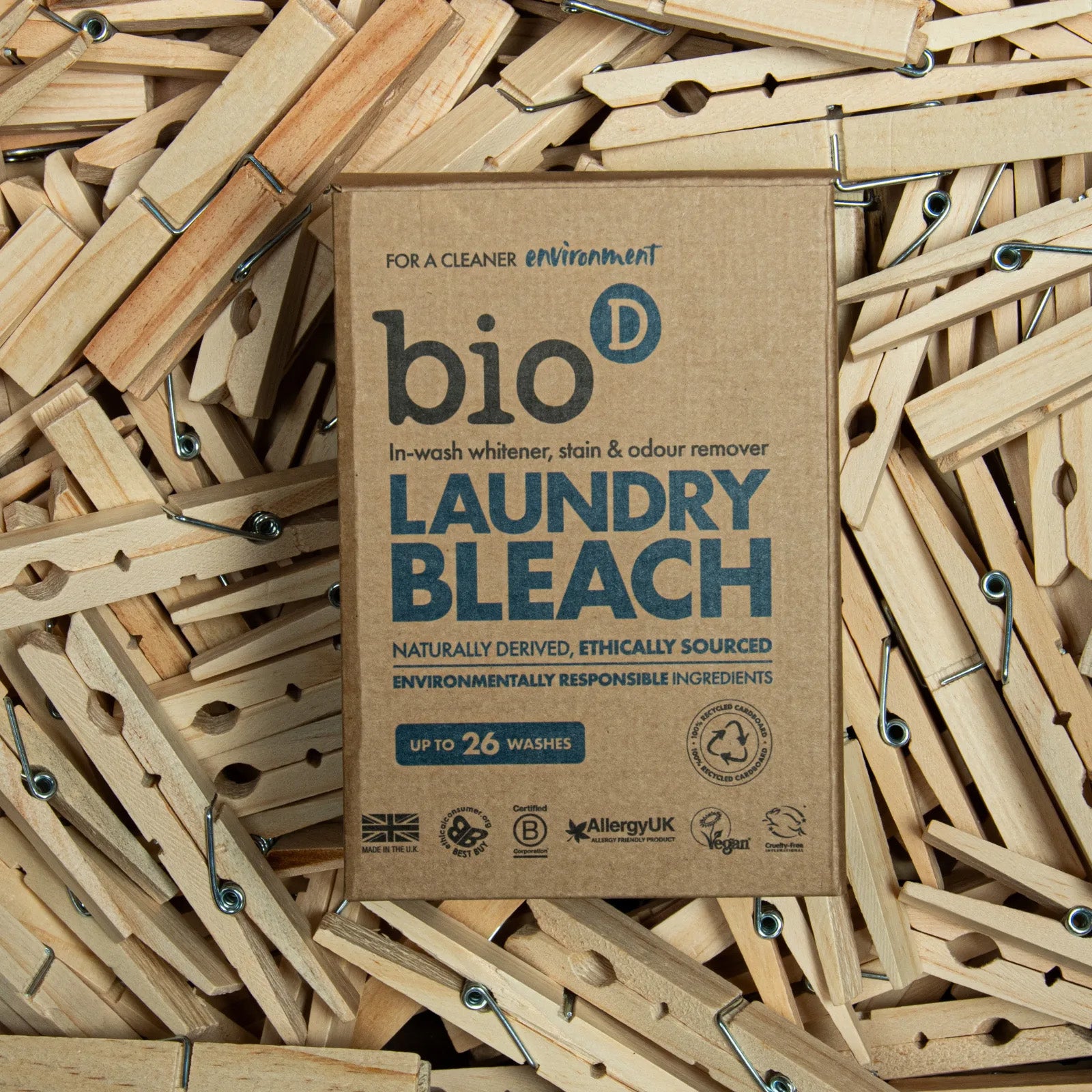 Front view of the Bio-D Laundry Bleach cardboard box placed on a background of wooden pegs, showing the product name, in-wash whitener description, and ethical certification icons.
