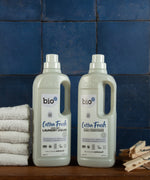 A 1L bottle of Bio-D cotton fresh laundry liquid and fabric conditioner standing on a wooden surface next to a pile of white towels and pegs