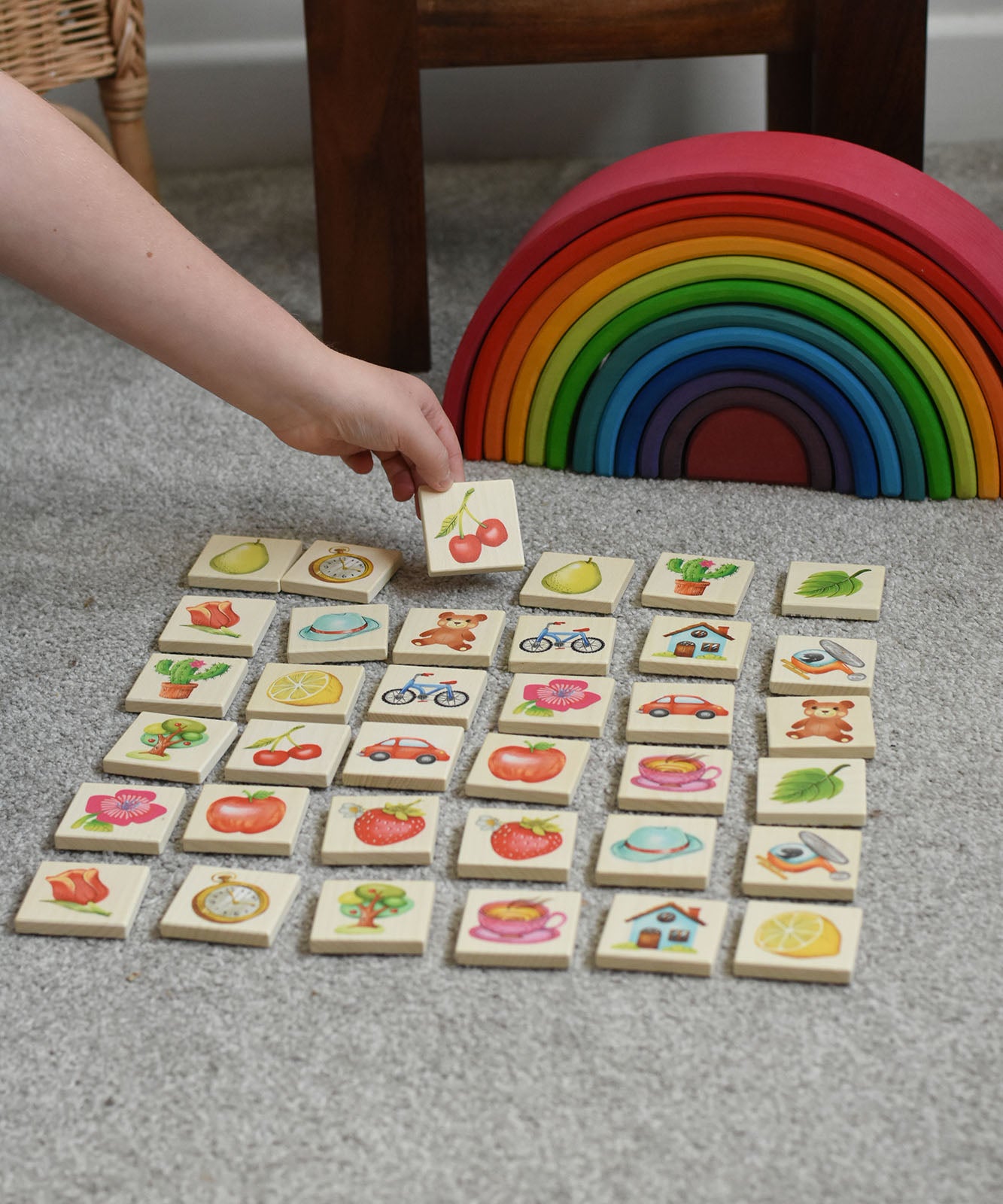 A child picking a tile from the Bajo Wooden Tiles Memory Game. The tile has an illustration of a cherry on it. All the tiles from the set have been laid out and are showing the different illustration. These heirloom quality toys by Bajo are part of a wide range of wooden toys available here at Babipur. 