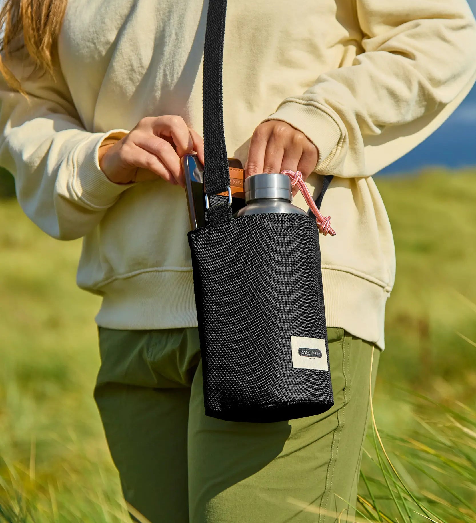 Adult carrying a Black + Blum insulated  bottle bag in black over the shoulder showing a flask and mobile phone inside