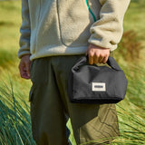 Adult carrying a Black+Blum 7 litres roll top lunch bag in black and standing in grass