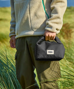 Adult carrying a Black+Blum 7 litres roll top lunch bag in black and standing in grass