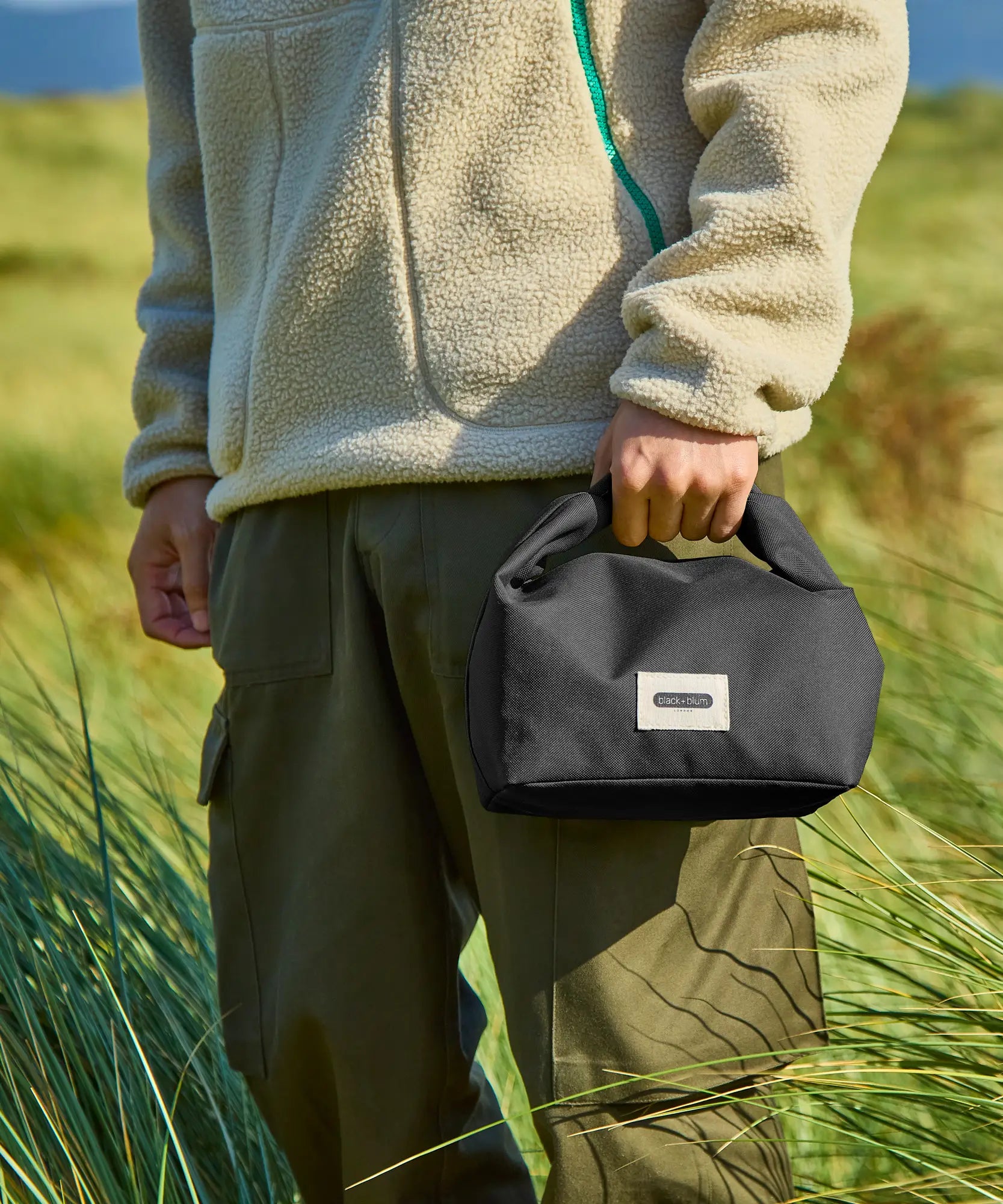 Adult carrying a Black+Blum 7 litres roll top lunch bag in black and standing in grass