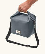 Adult hand carrying the Black+Blum 10 litres insulated lunch bag in slate grey by the roll top handle