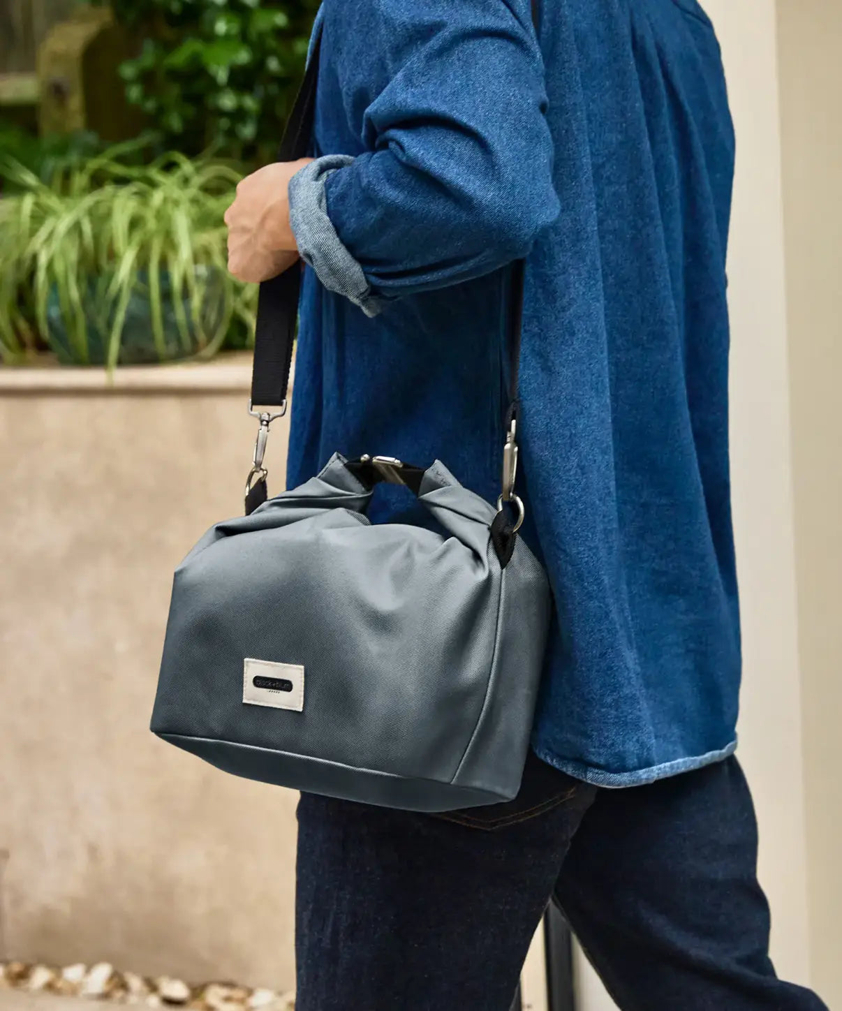 Adult carrying the Black+Blum 10 litres insulated roll top lunch bag in slate blue  over the shoulder with the carry strap