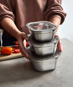 A adult standing by a stone worktop with the 3 different sized Black + Blum stainless steel lunchbox stacked on top of eachother