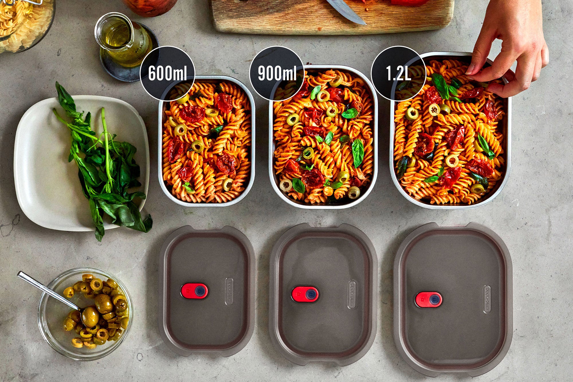 A top down view of the 3 diferent sized Black + Blum stainless steel lunch boxes next to eachother on a stone worktop with food inside and the grey lids next to each lunchbox