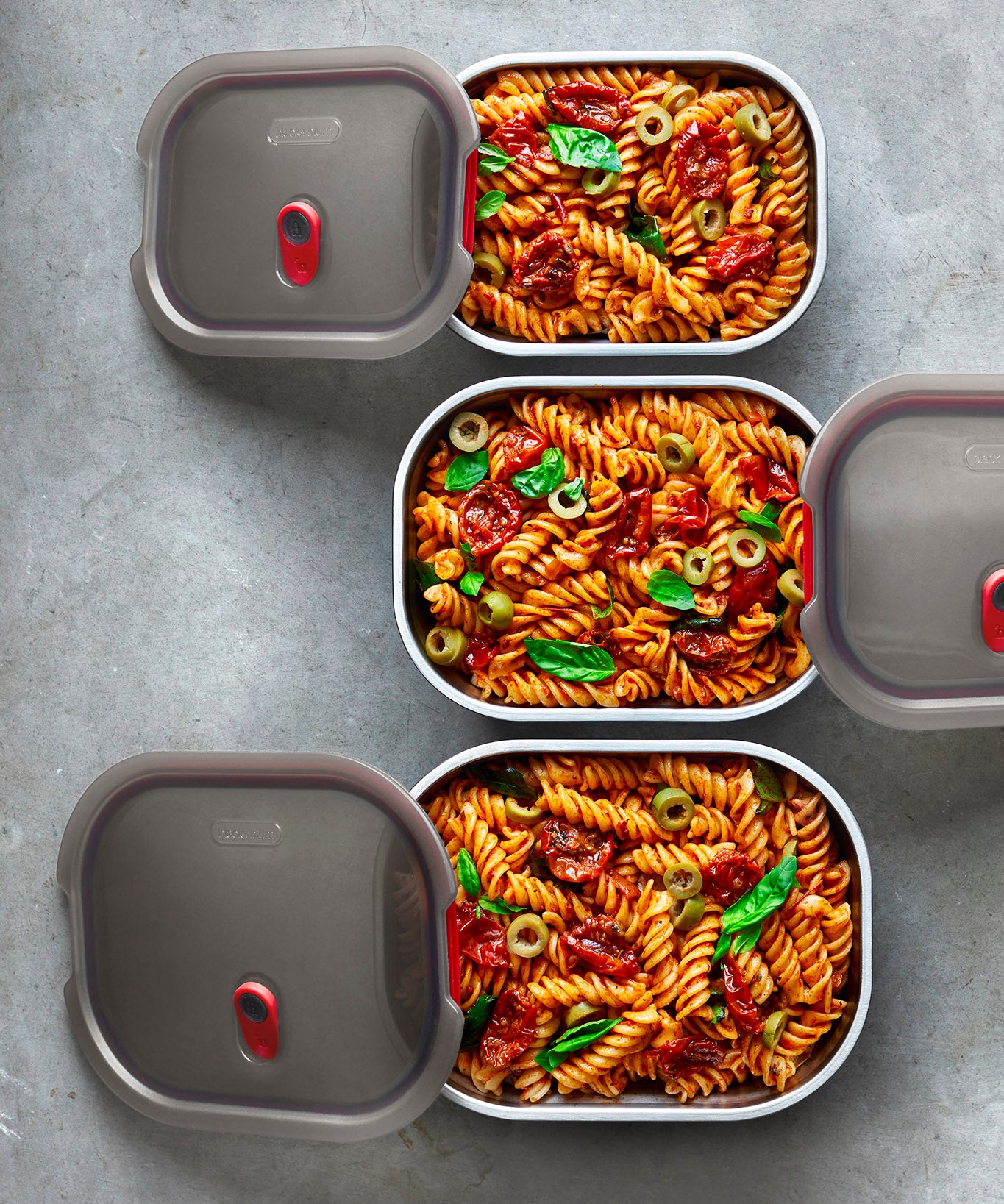 A top down view of the 3 sized Black + Blum stainless steel lunch boxes next to eachother on a stone worktop with food inside and the grey lids resting on top