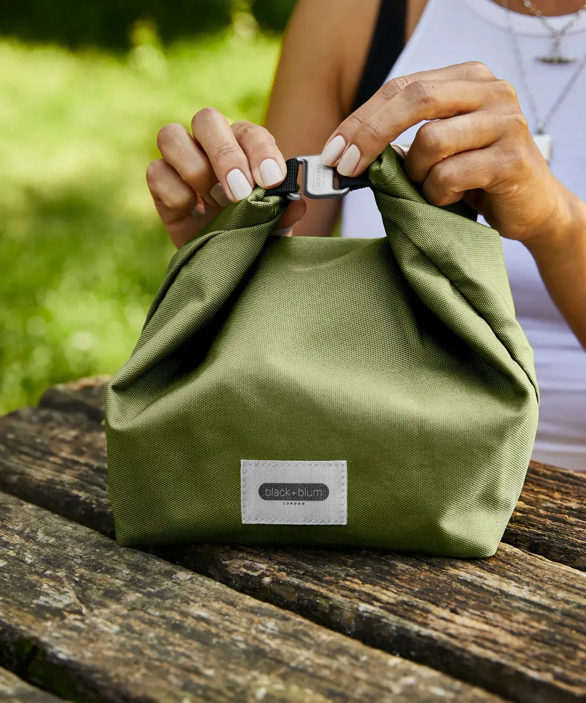 Adult closing a moss green Black+Blum 7 litres lunch bag using the hook buckle on a wooden table