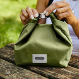 Adult closing a moss green Black+Blum 7 litres lunch bag using the hook buckle on a wooden table