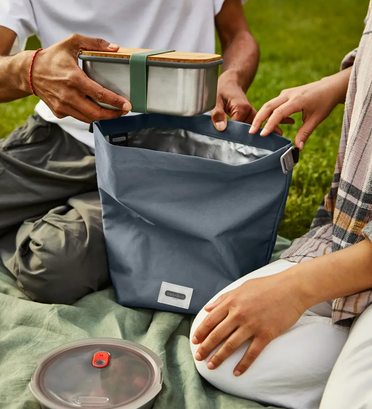 Adults packing a lunchbox in a slate blue Black+Blum 7 litres roll top lunch bag in black on a picnic blanket