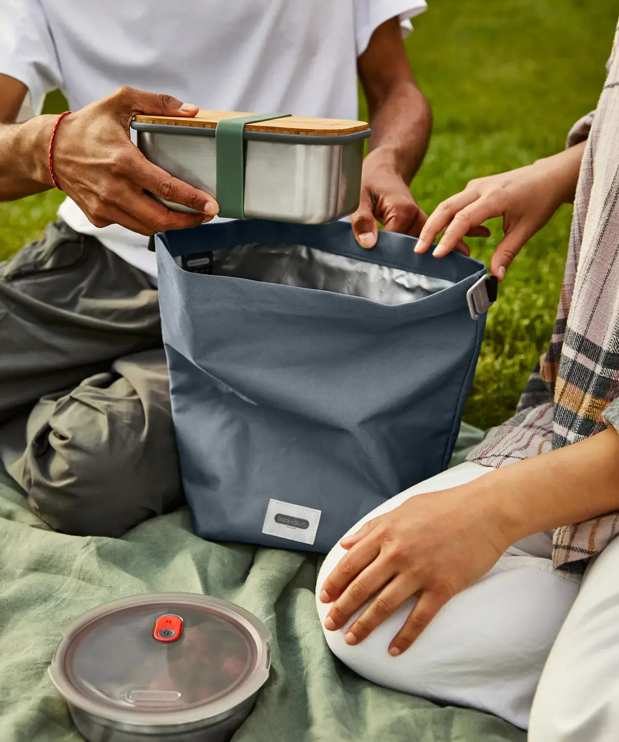 Adults packing a lunchbox in a slate blue Black+Blum 7 litres roll top lunch bag in black on a picnic blanket