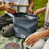 Adults packing a lunchbox in a slate blue Black+Blum 7 litres roll top lunch bag in black on a picnic blanket