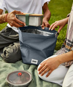 Adults packing a lunchbox in a slate blue Black+Blum 7 litres roll top lunch bag in black on a picnic blanket
