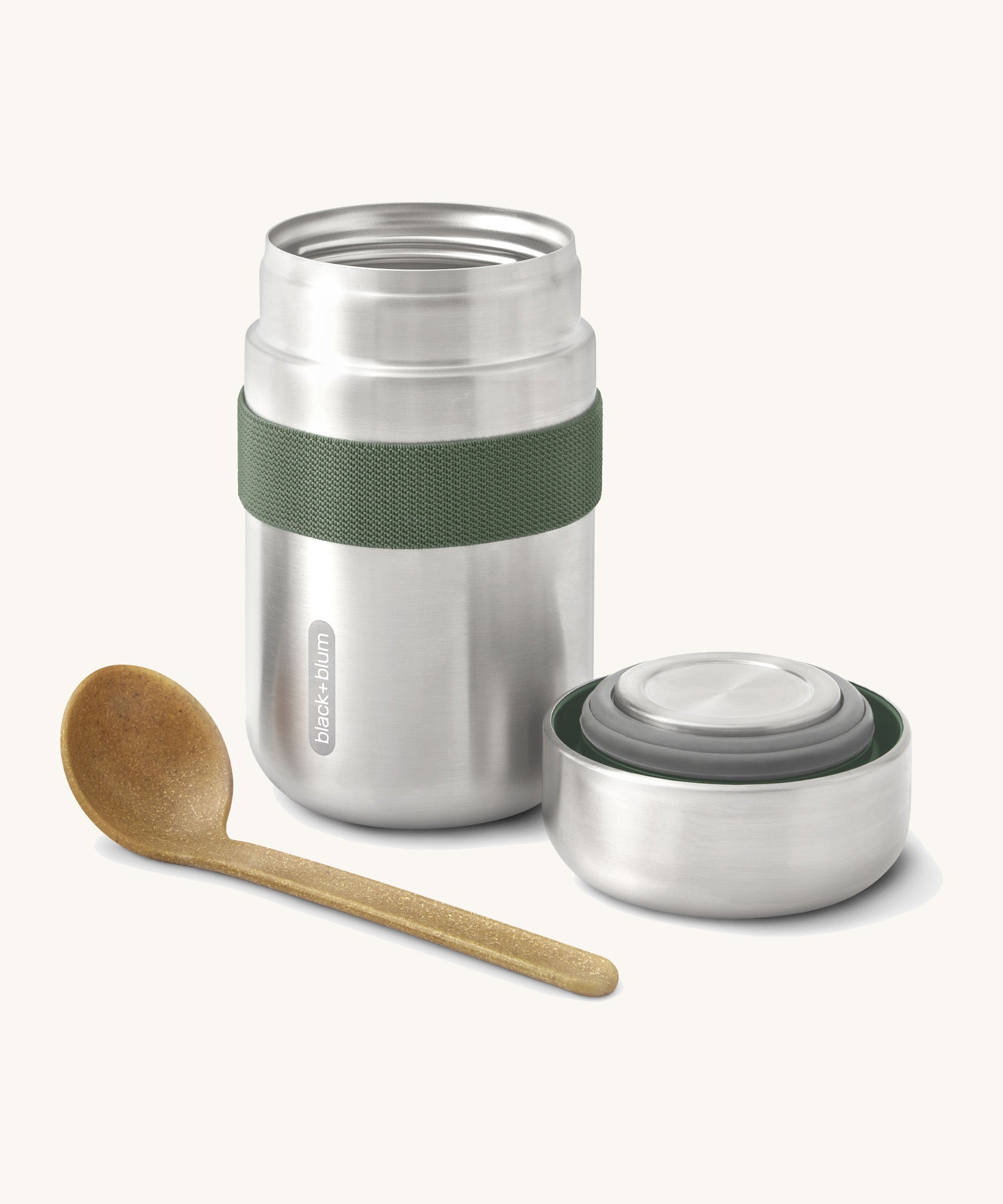 A close up of the Black + Blum insulated stainless steel food flask in olive showing the wooden spoon and lid resting next to the flask