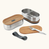 An open Black+Blum stainless steel leakproof bento lunchbox showing a fork and bamboo lid/chopping board