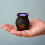 A Bumbu Wooden Witch's Cauldron. The cauldron is black on the outside and decorated with red, yellow and orange painted flames on the side. The top of the cauldron has a purple swirled effect decoration. The cauldron is resting in the palm of an adult's hand. 
