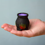 A Bumbu Wooden Witch's Cauldron. The cauldron is black on the outside and decorated with red, yellow and orange painted flames on the side. The top of the cauldron has a purple swirled effect decoration. The cauldron is resting in the palm of an adult's hand. 