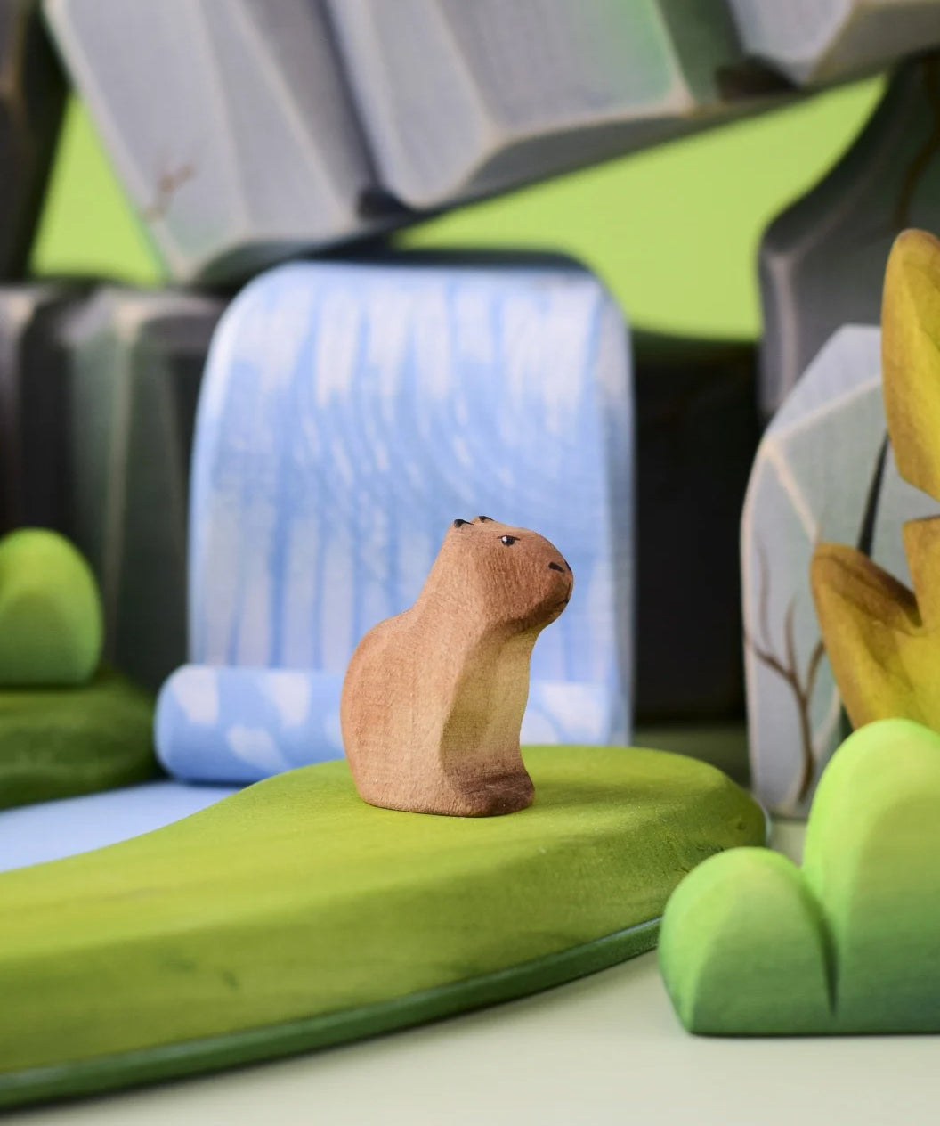 Bumbu Toys Wooden Sitting Baby Capybara in a scene with Bumbu rocks and grass.
