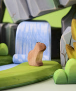 Bumbu Toys Wooden Sitting Baby Capybara in a scene with Bumbu rocks and grass.
