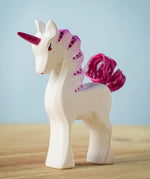 A Bumbu Toys Pastel Baby Unicorn wooden figure placed on a wooden surface. The figure is handmade and hand painted with pink coloured details