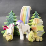 A close up of a Bumbu Toys Pastel Baby Unicorn wooden figure in a small world play scene with rainbow rocks and waterfall in the background. 