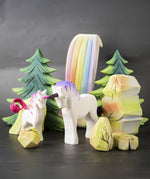 A close up of a Bumbu Toys Pastel Baby Unicorn wooden figure in a small world play scene with rainbow rocks and waterfall in the background. 