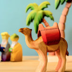 Bumbu wooden camel with red saddle pictured in a small world play scene with three kings and Bumbu palm trees in the background.