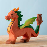 A Bumbu Toys handmade wooden dragon figure placed on a wooden surface. The dragon is red with green coloured wings and mane and orange coloured horns and chest. 
The dragon is part of a large collection of handmade wooden toy animals and scenery pieces that are available here at Babipur. 