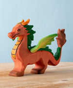 A Bumbu Toys handmade wooden dragon figure placed on a wooden surface. The dragon is red with green coloured wings and mane and orange coloured horns and chest. 
The dragon is part of a large collection of handmade wooden toy animals and scenery pieces that are available here at Babipur. 