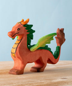 A Bumbu Toys handmade wooden dragon figure placed on a wooden surface. The dragon is red with green coloured wings and mane and orange coloured horns and chest. 
The dragon is part of a large collection of handmade wooden toy animals and scenery pieces that are available here at Babipur. 