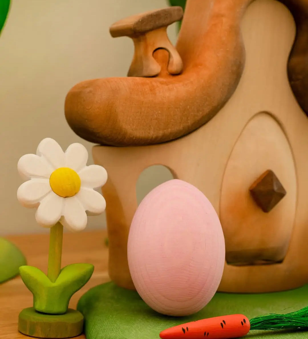 Bumbu handcrafted wooden pink egg resting infront of a wooden house, white daisy and carrot figure