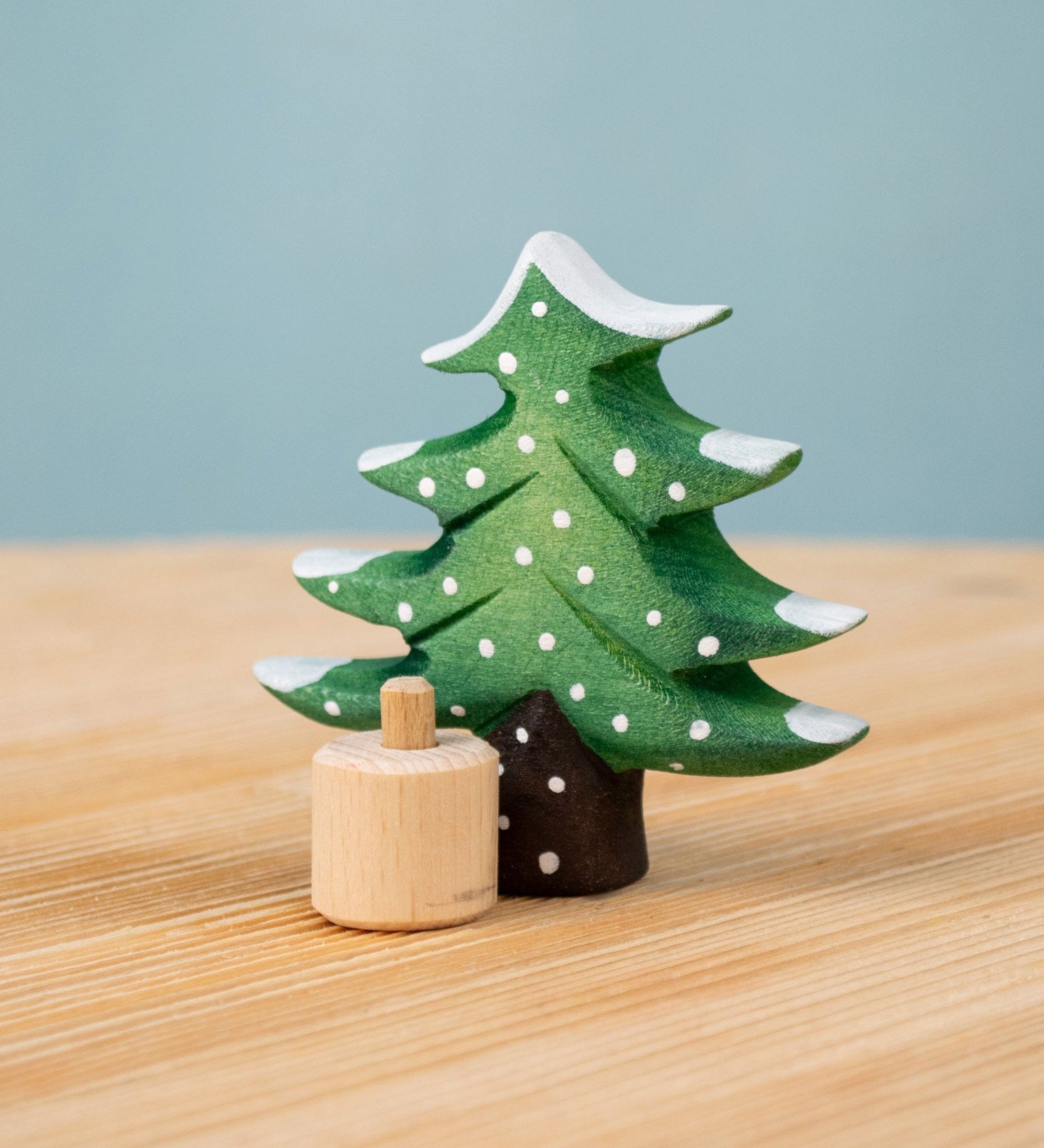 Bumbu Festive Tree Celebration decorative Figure to fit on a Grimm's celebration ring. The figure is a green fir tree decorated with snow. The wooden tree is shown separated from the wooden base it comes with.  These festive figures are perfect for your Christmas celebration ring set up. These expertly hand crafted items are part of a large range of wooden toys available here at Babipur. 