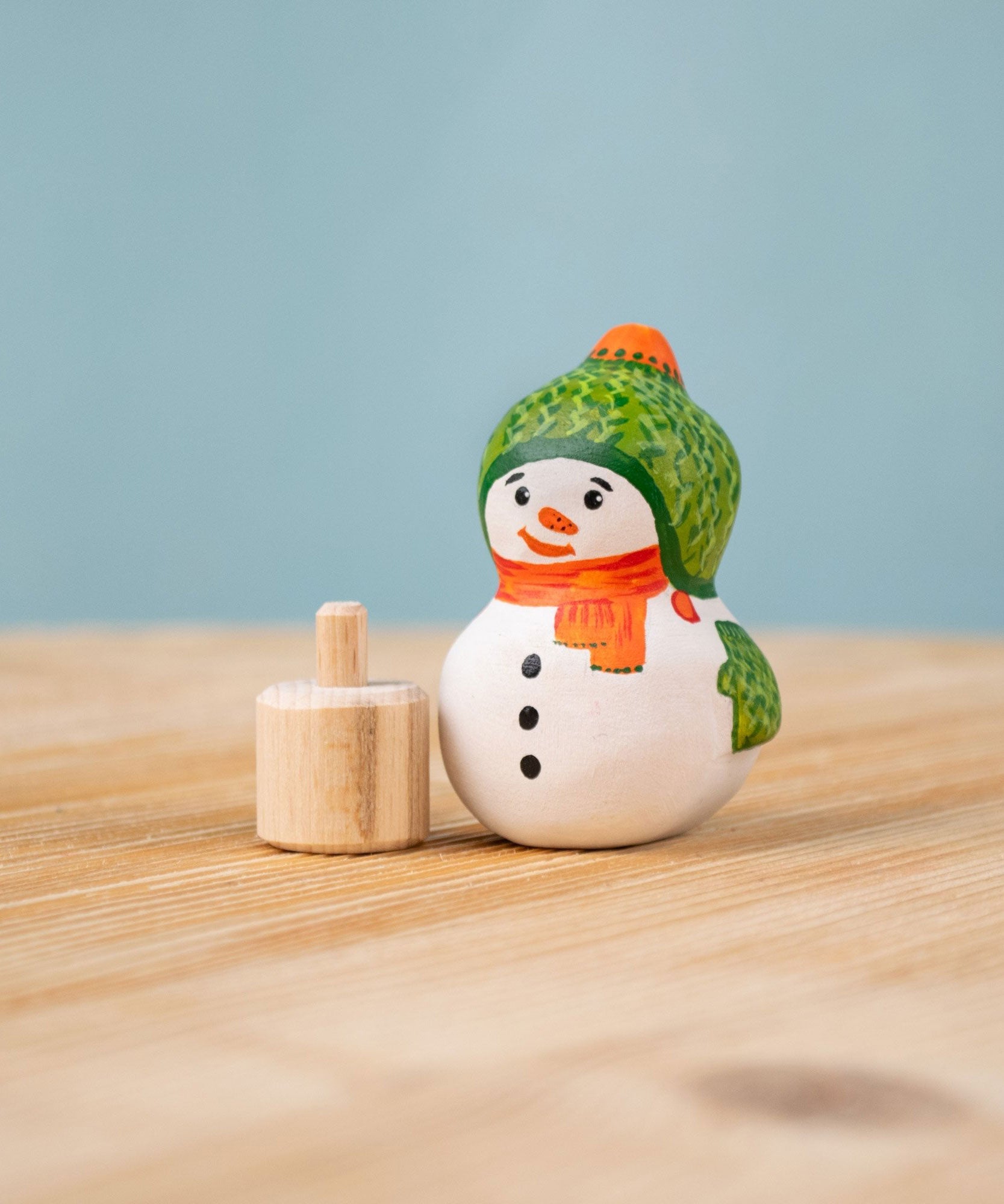 Bumbu Festive Snowman Celebration decorative Figure to fit on a Grimm's celebration ring. The hand painted snowman figure has a green hat with matching gloves and an orange scarf. The figure is shown separated from the wooden base that it comes with. These festive figures are perfect for your Christmas celebration ring set up. These expertly hand crafted items are part of a large range of wooden toys available here at Babipur. 
