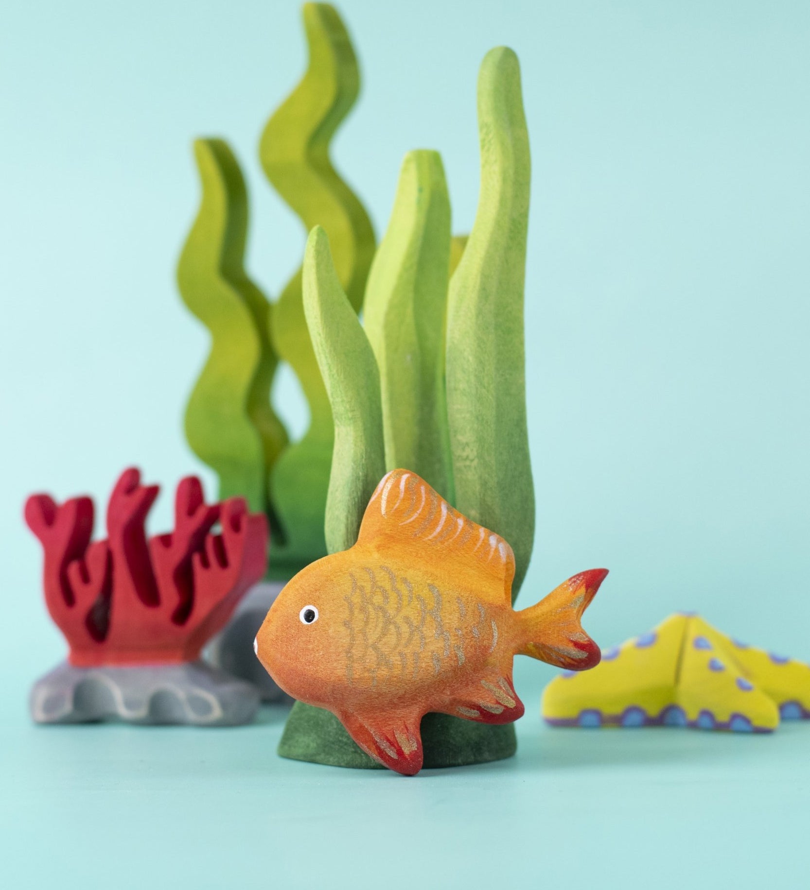The Bumbu hand made wooden goldfish toy figure, in an ocean playscene with seaweed and coral.