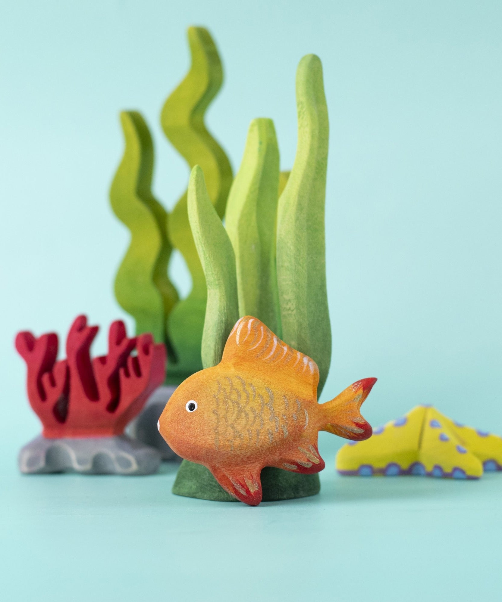 The Bumbu hand made wooden goldfish toy figure, in an ocean playscene with seaweed and coral.