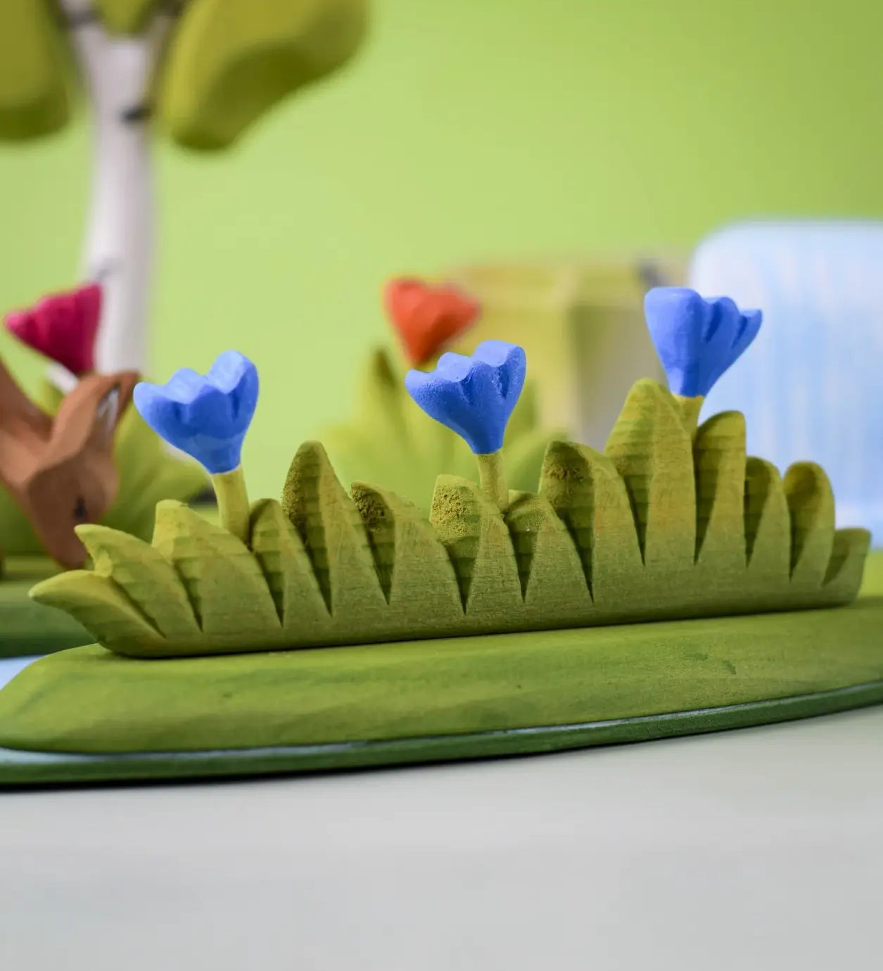 Bumbu large grass with 3 blue flowers wooden toy scenery piece placed at the front of a small world play scene. 