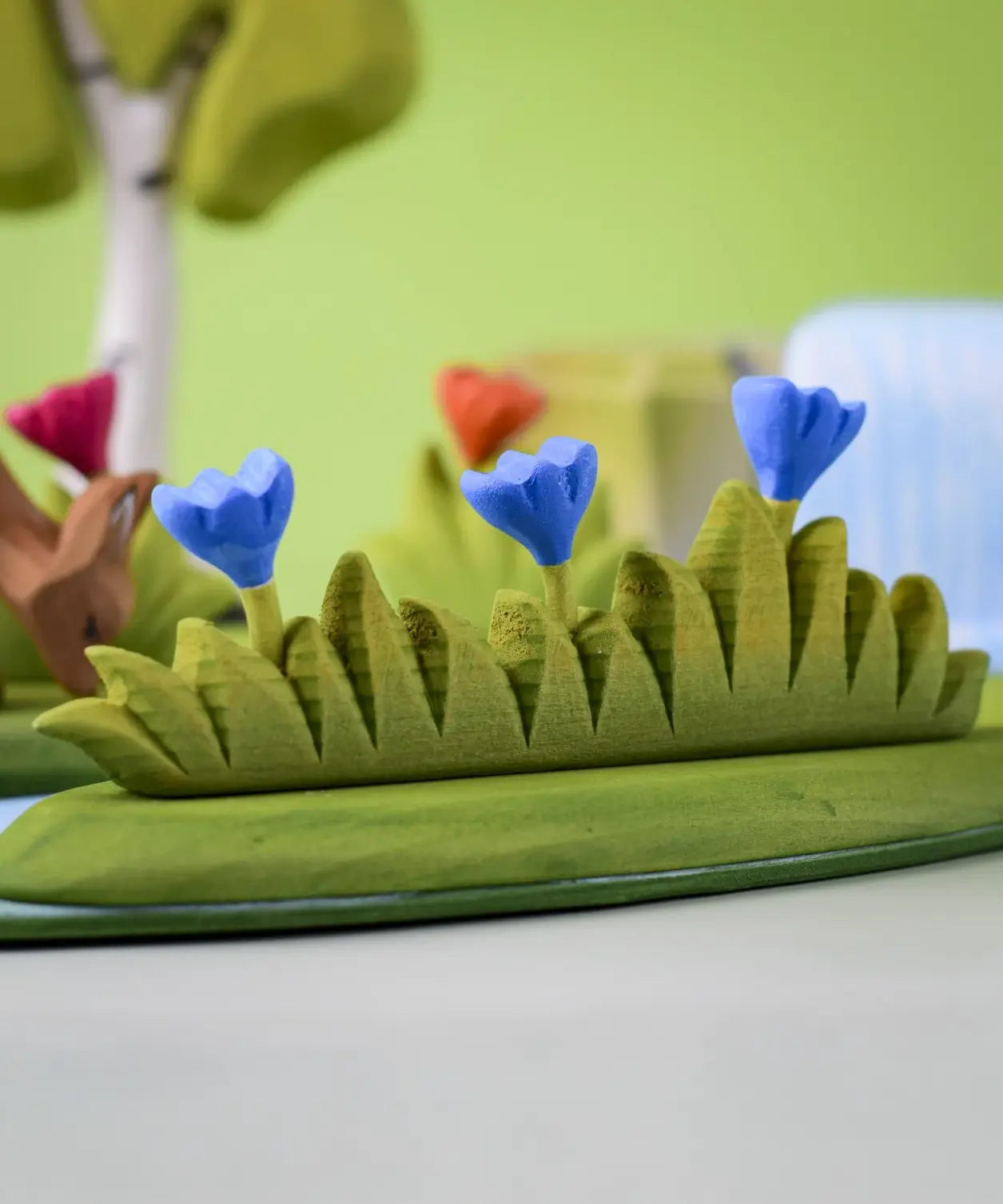 Bumbu large grass with 3 blue flowers wooden toy scenery piece placed at the front of a small world play scene. 
