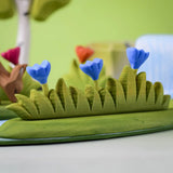 Bumbu large grass with 3 blue flowers wooden toy scenery piece placed at the front of a small world play scene. 