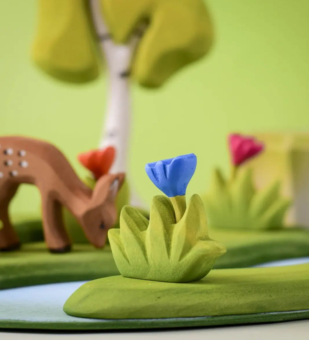 Bumbu small wooden grass with blue flower placed at the front of small world play scene with deer and trees in the background.