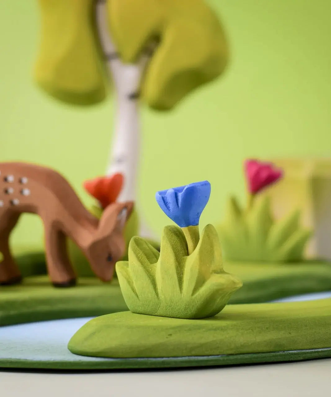 Bumbu small wooden grass with blue flower placed at the front of small world play scene with deer and trees in the background.