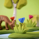 Bumbu small wooden grass with blue flower placed at the front of small world play scene with deer and trees in the background.