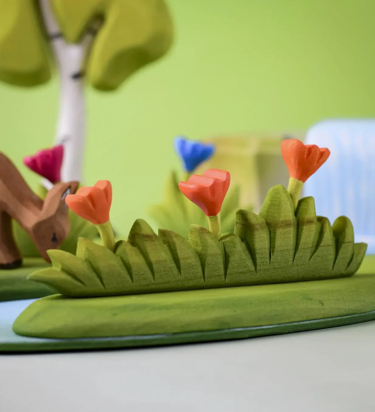 Bumbu large grass with three orange flowers wooden toy scenery placed at the front of a small world play scene with other flowers and trees in the background.
