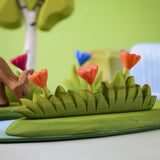 Bumbu large grass with three orange flowers wooden toy scenery placed at the front of a small world play scene with other flowers and trees in the background.