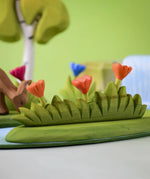 Bumbu large grass with three orange flowers wooden toy scenery placed at the front of a small world play scene with other flowers and trees in the background.