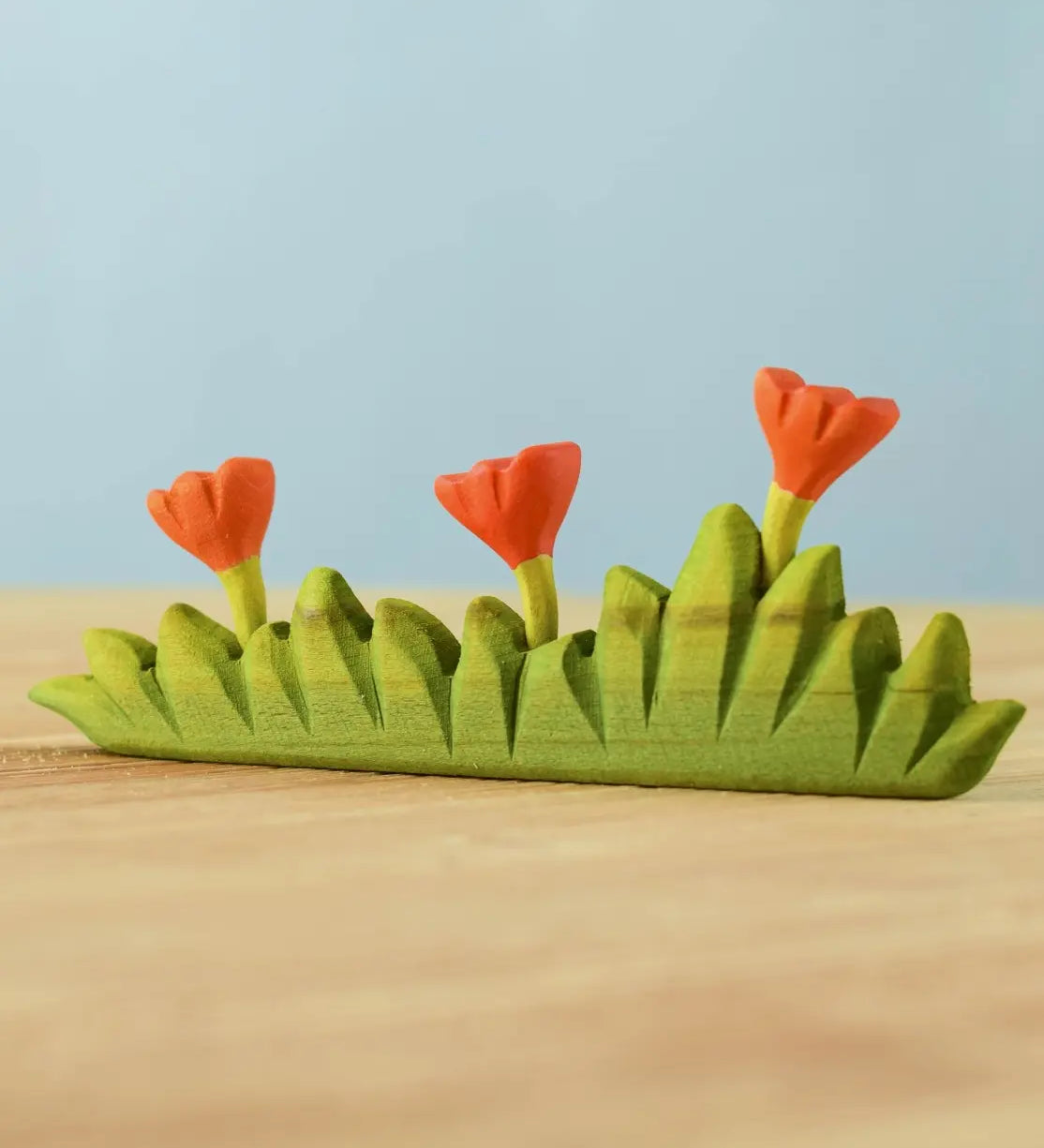 Bumbu large grass with three orange flowers wooden toy scenery placed on a wooden surface.
