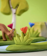 Bumbu small wooden grass with orange flower scenery piece placed in a small world play scene with deer, flowers and trees in the background.