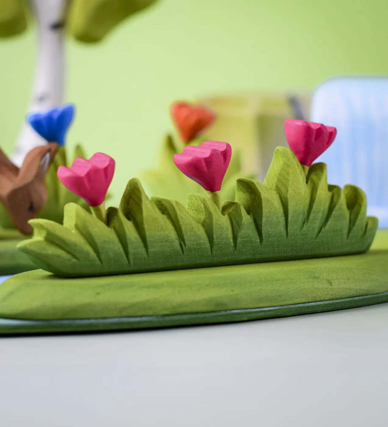 Bumbu large wooden grass with three pink flowers toy scenery piece placed at the front of a small world play scene with other flowers and trees in the background.