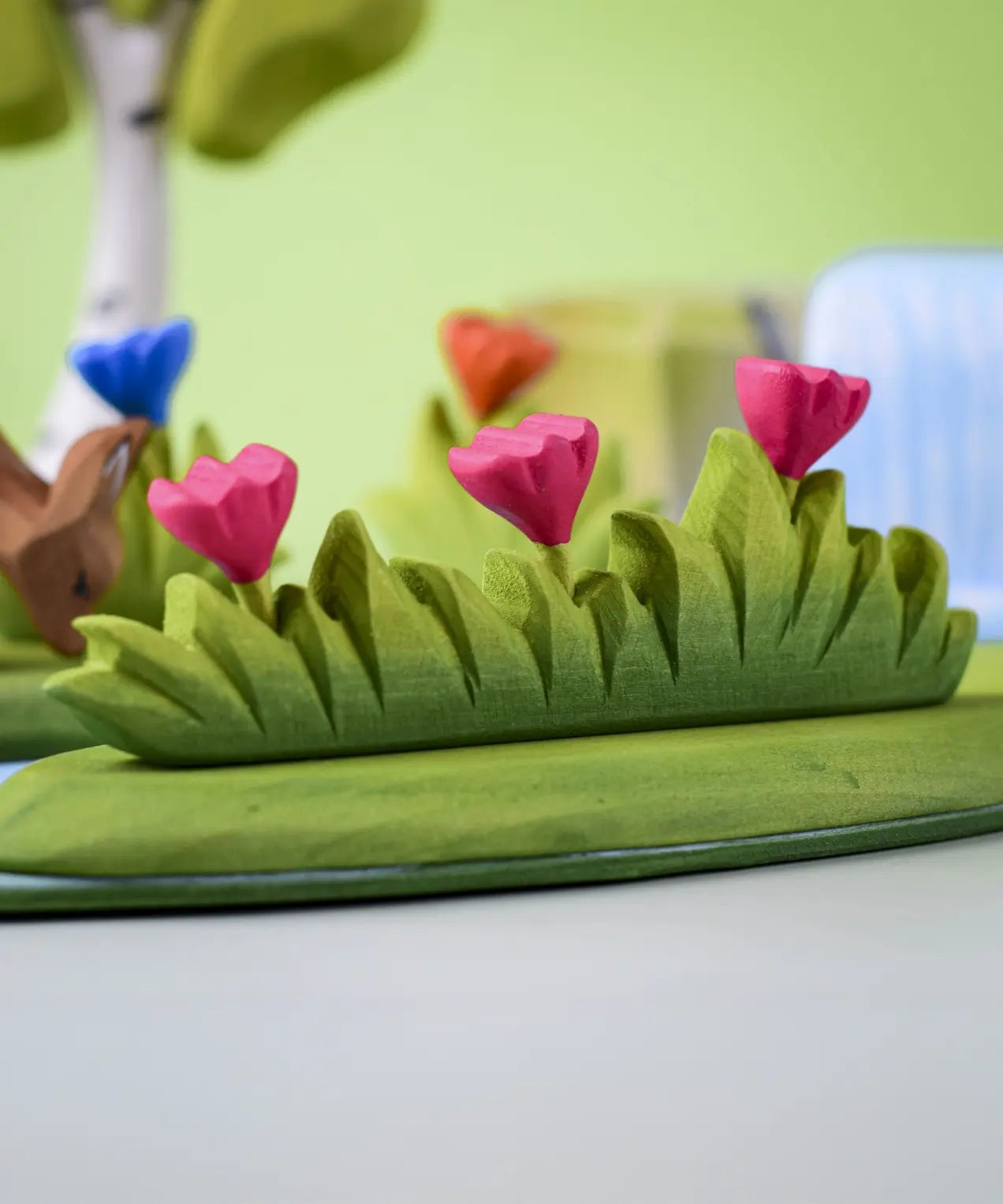 Bumbu large wooden grass with three pink flowers toy scenery piece placed at the front of a small world play scene with other flowers and trees in the background.