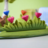 Bumbu large wooden grass with three pink flowers toy scenery piece placed at the front of a small world play scene with other flowers and trees in the background.